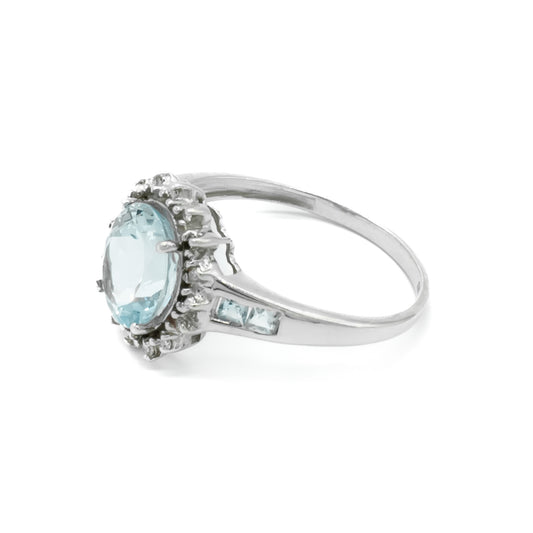 Classic 9ct white gold ring set with a beautifully faceted, light blue aquamarine surrounded by twelve tiny diamonds and two princess cut aquamarines on each shoulder.